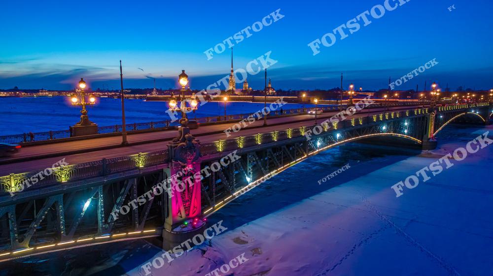 Фотография зимнего Санкт-Петербурга с высоты дрона с панорамным видом на Троицкий мост, Петропавловскую крепость и реку Неву с ледяной поверхностью. На снимке видны исторические здания, мосты и набережные, освещенные вечерними огнями, создавая атмосферу романтики и величия северной столицы России. Панорамный ракурс позволяет оценить масштабы городской архитектуры и транспортных сооружений, подчеркивая гармонию природы и урбанистики Петербурга. Изображение идеально подходит для туристических публикаций, зимних коллекций фотографий, постеров, интерьерного оформления и презентаций о городской архитектуре.